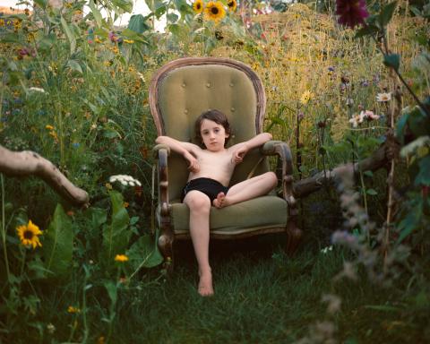 Visuel The Garden, un enfant sur un fautueil vert, fixe la caméra et est entouré de verdure, haute herbes et fleurs