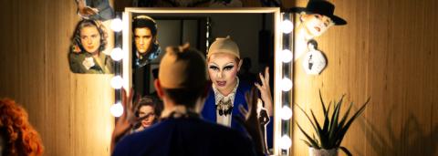 Une personne est assise de dos devant un miroir entouré d’ampoules allumées, dans une loge. Son reflet montre un visage transformé avec un maquillage drag très expressif, yeux soulignés de noir et lèvres accentuées. Elle porte une perruque beige sur la tête, en préparation d’une performance. Le miroir est décoré de découpages de portraits de stars (dont Elvis Presley et d'autres icônes hollywoodiennes)