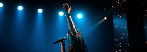 Photo d'une personne avec son micro, le bras levé et les projecteurs en arrière-plan