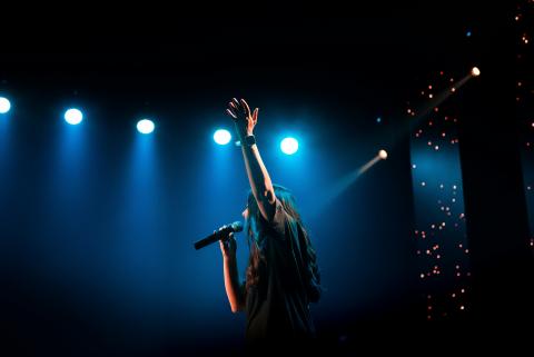 Une personne au cheveux long chante, un micro à la main et lève l'autre main, derrière on voit des lumière de scène bleu