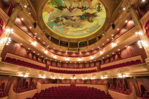 Intérieur d’un opéra à l’architecture classique, vu depuis la scène : plusieurs balcons ornés de dorures et de velours rouge entourent la salle, illuminés par de nombreux luminaires. Au plafond, une vaste fresque circulaire peinte représente des figures et des nuages dans des tons pastel